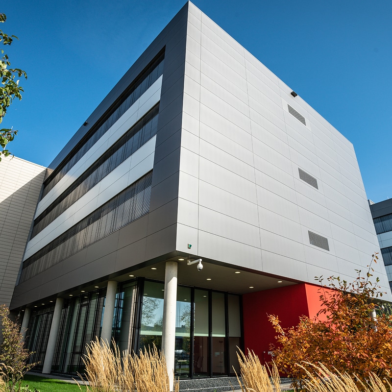 Bâtiment moderne : Façade élégante en verre et aluminium gris Bâtiment de bureaux moderne, angle en contre-plongée. Façade en panneaux gris argentés, entrée avec colonnes et mur rouge. Ciel bleu, herbes d'automne.