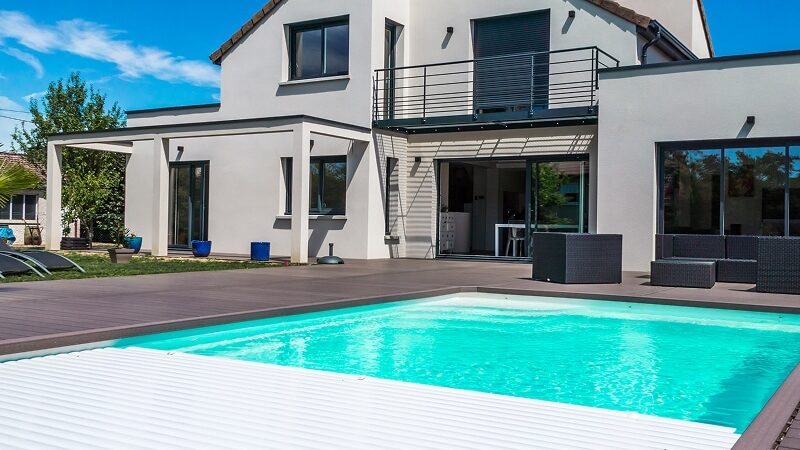 Architecture contemporaine, maison blanche et piscine avec volet roulant automatique blanc. Grande terrasse en bois foncé sous un ciel bleu.