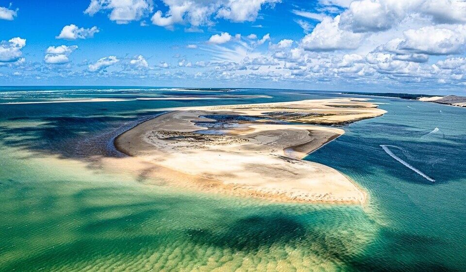 Banc de sable et eau turquoise : Splendeur près du Pilat Vue aérienne d'un grand banc de sable au bord d'un bassin. L'eau turquoise et émeraude contraste avec la Dune du Pilat au loin. Traînées de bateaux.
