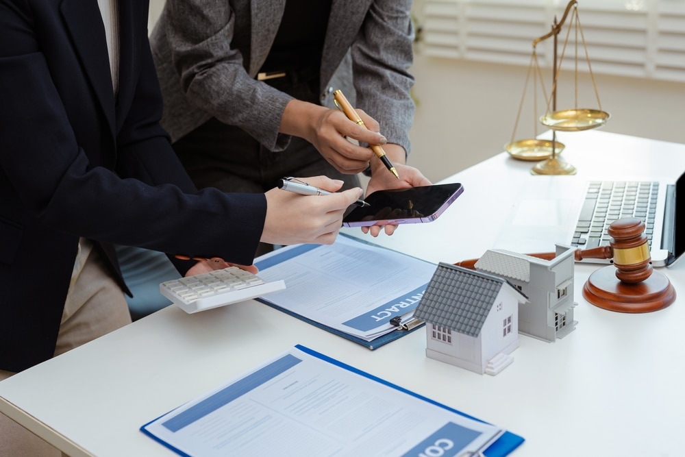 Contrat immobilier et achat maison : consultation légale. Deux professionnels analysent un contrat immobilier avec smartphone et calculatrice. Justice (balance, maillet) et maquettes de maisons sur le bureau.