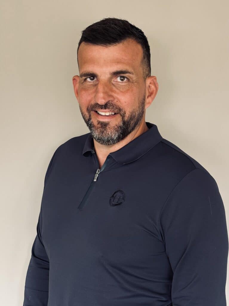 Portrait d'un homme souriant en polo bleu marine zippé Homme souriant barbu portant un polo bleu marine zippé devant un mur beige.