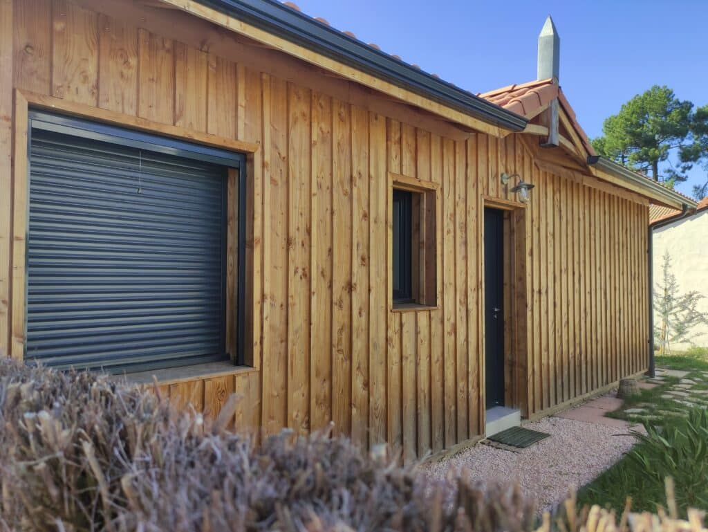 Maison en bois moderne : Bardage vertical et design épuré Façade de maison en bois avec bardage vertical, volet roulant gris anthracite et porte moderne sur jardin.